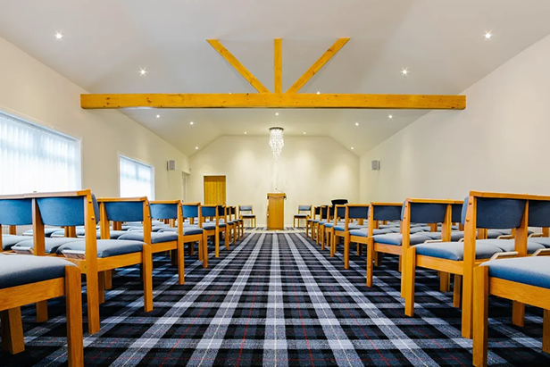 Chapel of Rest featuring a timber beamed ceiling, wooden chairs with blue cushions and a blue checked carpet.